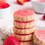 A stack of strawberry shortbread cookies with strawberry glaze on a white plate. A fresh strawberry is on the plate and a small bowl of strawberry glaze in the background