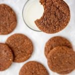 a cocoa molasses spice cookie balanced on the rim of a glass of milk with more cookies on the table below