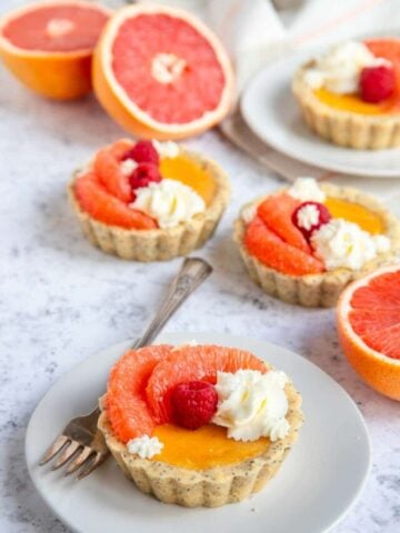 Grapefruit curd mini tarts decorated with whipped cream, fresh grapefruit and fresh raspberries. Fresh cut grapefruit sit next to the tarts