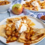 Apple cinnamon crepes on a white plate topped with whipped cream. More filled crepes in the bacground