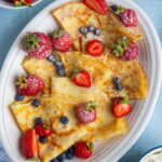 Platter with folded crepes, topped with strawberries, blueberries and dusted with powdered sugar