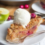 A slice of cranberry apple crumble pie on a dessert plate topped with ice cream. Another slice and the pie are in he background