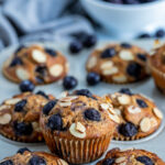 a wholte wheat blueberry muffin ssitting on tops of a muffin pan surrounded by other blueberry muffins