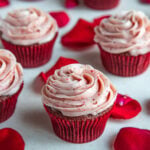 Frosted red velvet cupcake next to other cupcakes and surrounded by rose petals