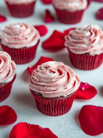 Frosted red velvet cupcake next to other cupcakes and surrounded by rose petals