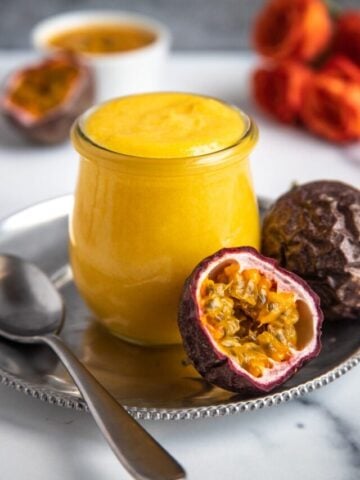 Passion fruit curd in a jar on a small platter next to fresh passion fruit and a serving spoon. Flowers and passion fruit puree in the background