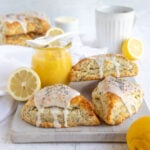 Lemon Poppy Seed Scones with lemon glaze on a cooling rack set in a small sheet pan. Whole lemons and half lemons next to the sheet pan with a small jar of lemon curd