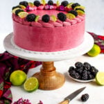 Blackberry lime cake on pedestal surrounded by fresh blackberries and fresh limes