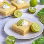Key lime bar on a dessert plate with a dollop of whipped cream. More bars in the background with fresh key limes