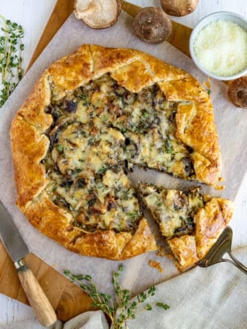 mushroom galette on a wooden serving board with a cutting knife, bowl of parmesan cheese and some fresh thyme