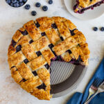 blueberry pie with one piece cut out. Another piece of pie is on a dessert plate north of the whole pie with a bowl of blueberries to the left