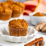 Pumpkin muffin on small saucer with cinnamon sticks and whole nutmeg next to the plate. Fresh ginger and more muffins are in the background