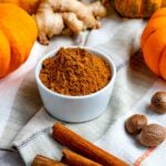 pumpkin pie spice in a white container surrounded by cinnamon sticks, whole nutmeg, fresh ginger and mini pumpkins
