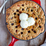 chocolate chip skillet cookie in a red Le Creuset cast iron skillet with three scoops of ice cream. Two spoons are on the left with a glass of milk in the upper right hand corner next to the skillet