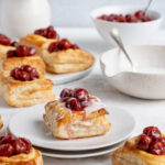 Red tart cherry cream cheese puff pastry on dessert plate. In the background there is a large plate with 4 pastries. On the right side is a bowl of lemon glaze and another bowl with the cherry filling.