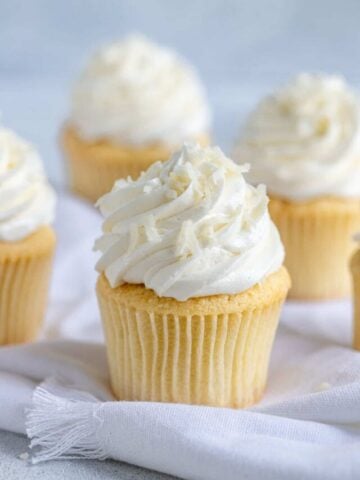 Coconut cupcake on a white table linen with more frosted cupcakes behind it.