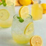 2 glasses of lemon soda. Each glass contains lemon simple syrup, club soda, ice cubes, and mint leaves. There are fresh lemons in the background