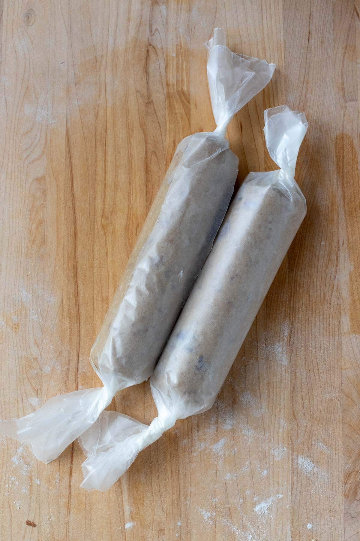 The brown sugar pecan cookie dough shaped into two logs and wrapped in wax paper. The cookie dough logs are on a wooden work surface