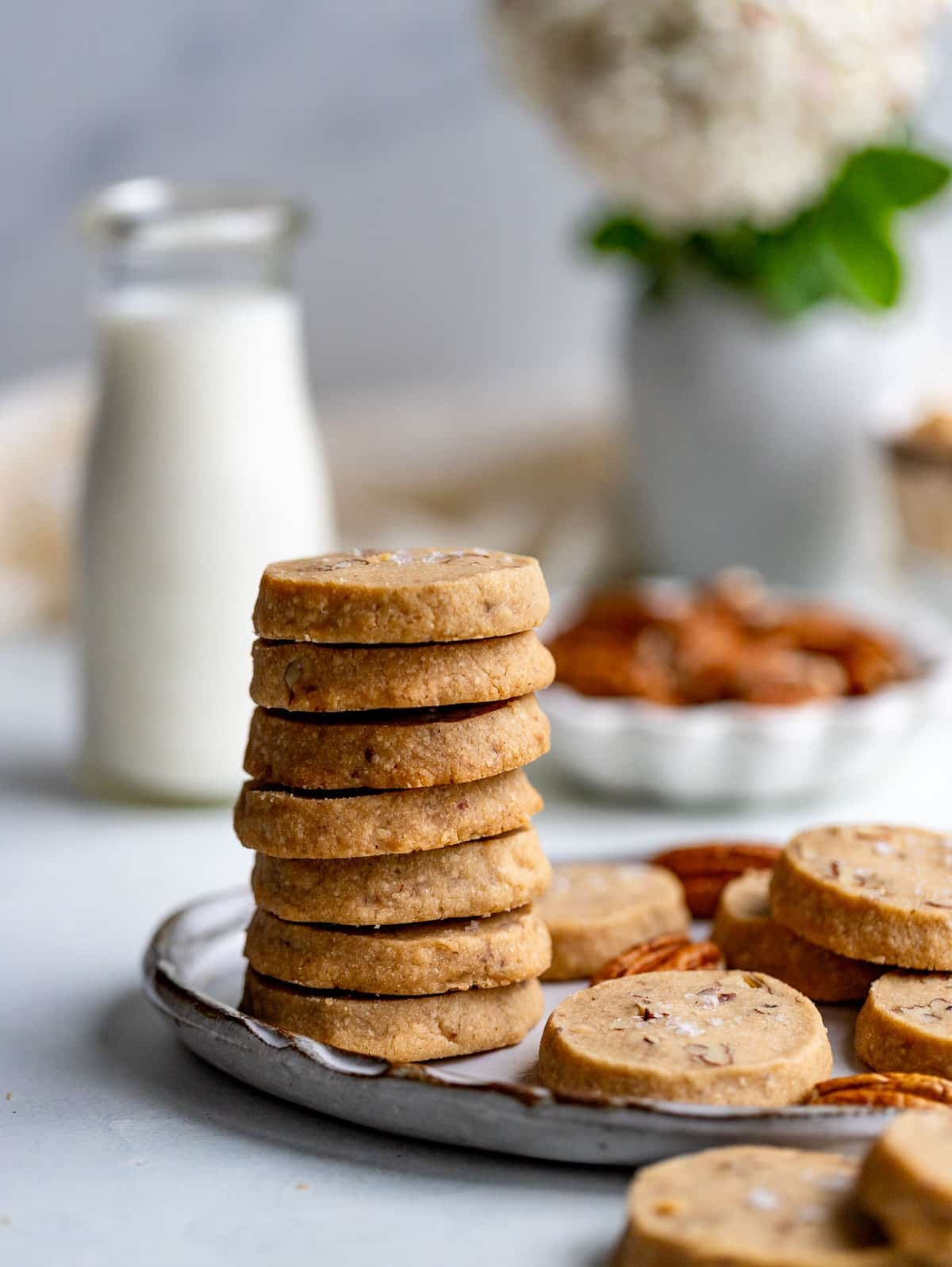 A stack of 7 brown sugar pecan cookies on a light gray plate with more cookies in a single layer next to the stack. Behind the stack of cookies is a class of milk, a small bowl of pecans and bud vase with a single flower