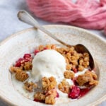 A serving of rhubarb crumble in a cream colored ceramic bowl with a scoop of vanilla ice cream on top. In the background there is baking dish with the rhubarb crumble and another serving of rhubarb crumble