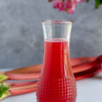 Rhubarb syrup in a glass jar with long stalks of fresh rhubarb in the background and a small vase with pink flowers