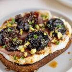 A close up image of ricotta toast on a small plate. The ricotta toast is made with toasted bread topped with ricotta, prosciutto, prunes, chopped pistachios, fresh herbs, and a honey balsamic glaze.