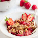 Strawberry crumble in a white bowl with a spoon. There are 3 fresh half strawberries on top of the crumble. Behind the bowl is a decorative ceramic bowl with fresh strawberries and 3 fresh strawberries on the table next to the ceramic bowl