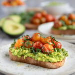 Avocado toast with tomato on a light green salad plate. An avocado cut in half, another serving of the avocado toast, fresh cherry tomatoes, and a bowl of roasted cherry tomatoes are in the background.