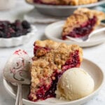 A slice of marionberry pie on a dessert plate with a scoop of vanilla ice cream next to the pie. Another slice of pie and the remaining pie is in the background