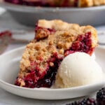 A slice of marionberry pie on a dessert plate with a scoop of vanilla ice cream next to the pie. Another slice of pie and the remaining pie is in the background