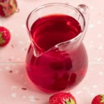 Strawberry simple syrup in a small glass container with a handle and a pour spout. The container is on a pick surface with red and white dots an surrounded by several fresh strawberries