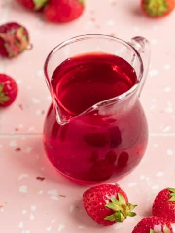 Strawberry simple syrup in a small glass container with a handle and a pour spout. The container is on a pick surface with red and white dots an surrounded by several fresh strawberries