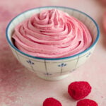 A bowl of raspberry buttercream piped into a blue and white ceramic bowl. There are fresh raspberries next the bowl