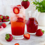 Strawberry soda in a 12-ounce glass with fresh sliced strawberries added to the soda. A small container of strawberry simple syrup is next to the glass of homemade soda. A bowl of fresh strawberries is behind the drink to the left. A small vase with fresh flowers is behind and to the right of the homemade soda.
