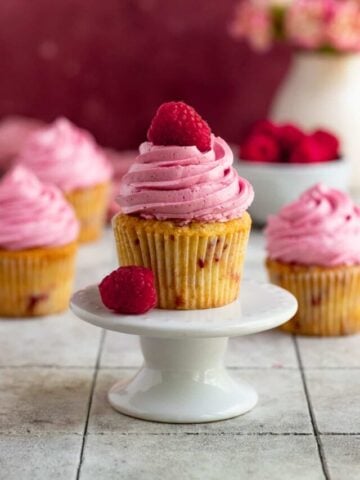 A single raspberry cupcake with raspberry buttercream on a mini cake stand. The cupcake is topped with a fresh raspberry. There are three cupcakes in the background and a small bowl of fresh raspberries.