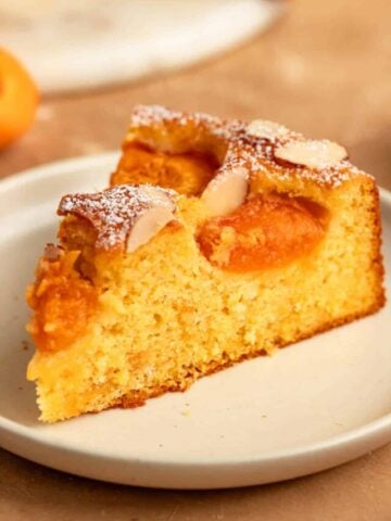 A slice of apricot cake on a dessert plate. The cake is topped with apricot halves, sliced almonds, and a dusting of powdered sugar.