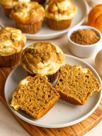 Two pumpkin muffins arranged on a small plate. One muffin is cut in half and the other muffin is whole. There is a large plate of muffins behind and to the right of this small plate