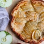 Apple puff pastry galette on a piece of parchment paper and surrounded by whole and cut Granny Smith apples