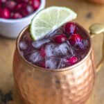 Cranberry moscow mule in a copper mug with a lime slice on top of the drink. The drink is filled with ice, and fresh cranberries