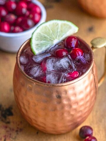 Cranberry moscow mule in a copper mug with a lime slice on top of the drink. The drink is filled with ice, and fresh cranberries