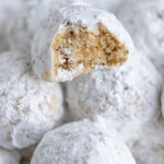 Pecan snowball cookies piled on a plate. The cookie on top has a piece taken out so you can see the light brown interior and pieces of pecans