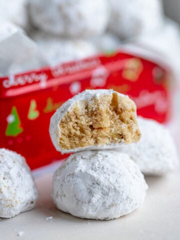 Four pecan snowball cookies arranged on a flat surface with one cookie on top of the others with a bite taken out of it. There is a red tin full of cookies behind the cookies