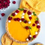 Mango curd tart with a coconut tart crust. The top of the tart is decorated with fresh mango and raspberries. There is a bowl of fresh raspberries to the left and a small bowl of sliced mangos to the right.