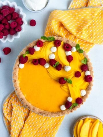 Mango curd tart with a coconut tart crust. The top of the tart is decorated with fresh mango and raspberries. There is a bowl of fresh raspberries to the left and a small bowl of sliced mangos to the right.