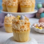 Coconut Easter Cupcake on top of a small pedestal. There is cake stand in the background with cupcakes.