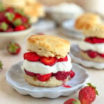 Strawberry shortcake on a small plate with a bowl of strawberries and another strawberry shortcake in the background.