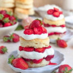 Strawberry shortcake on a small plate with a bowl of strawberries and another strawberry shortcake in the background.