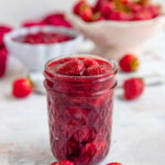 Roasted strawberries in an 8-ounce glass jar with more roasted strawberries in a bowl in the background
