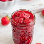 A pint jar filled with roasted strawberries. the strawberries are bright red. There is a small bowl of roasted strawberries in the background.