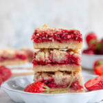 A stack of 3 strawberry crumb bars on a small white plate. Fresh strawberries and more bars in the background